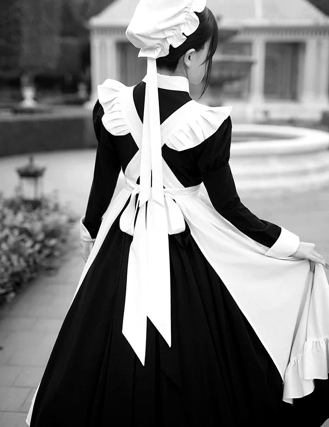 A black maid uniform with a white apron and cap, worn in a formal garden, seen from behind